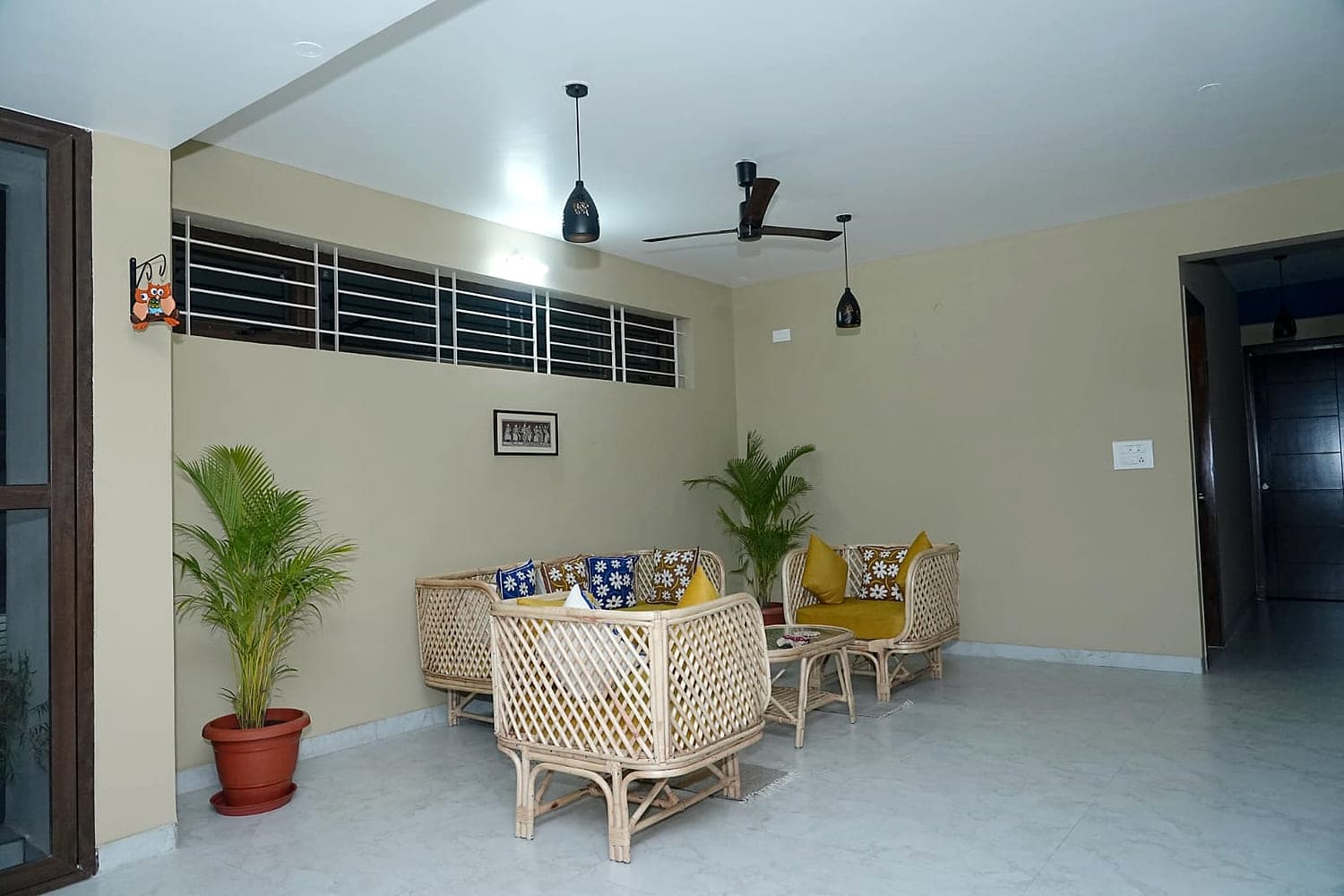 A wide living room with cane chairs, potted palms and a plain painted wall.