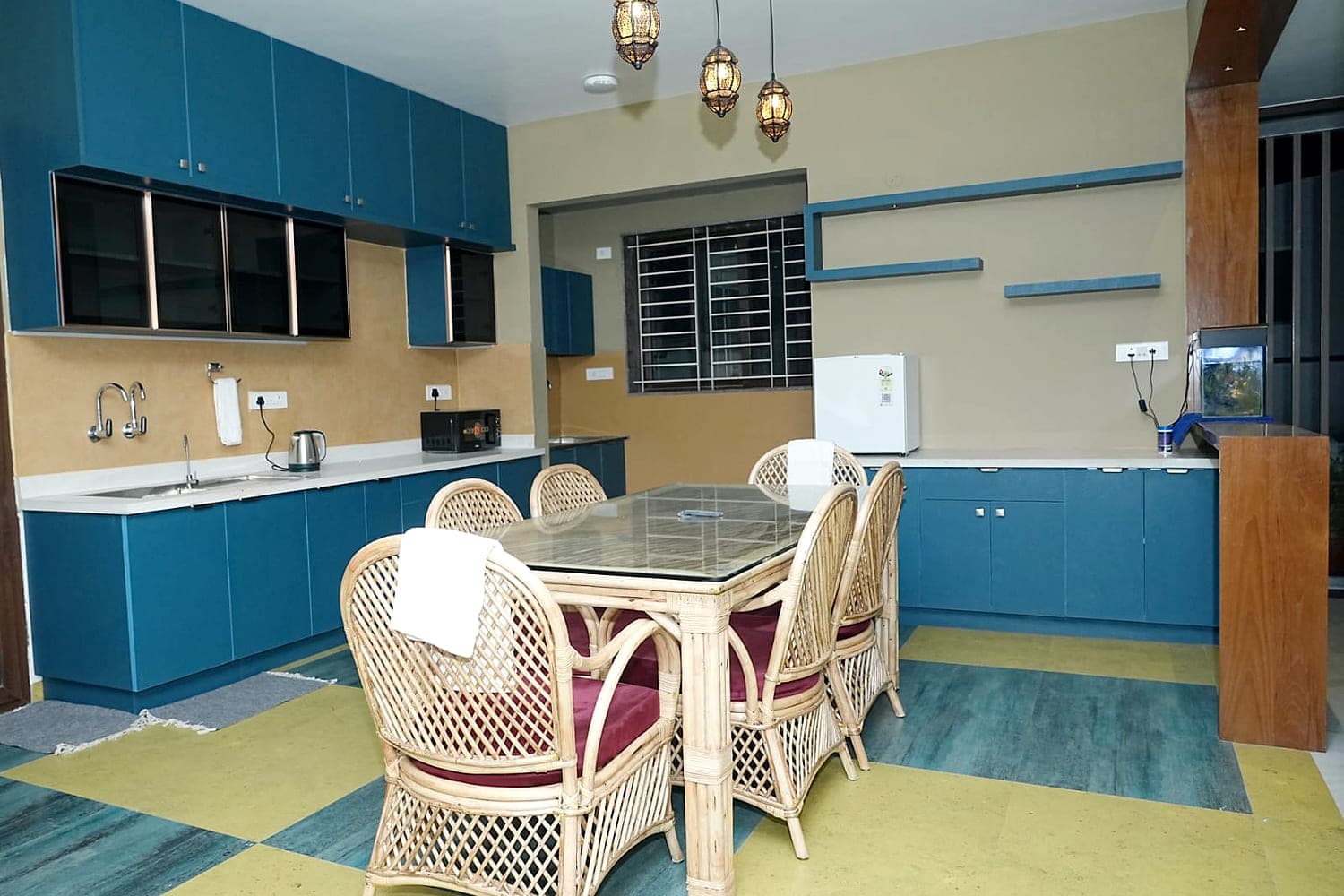 A blue kitchen with a cane dining set, a kettle and a small fridge.