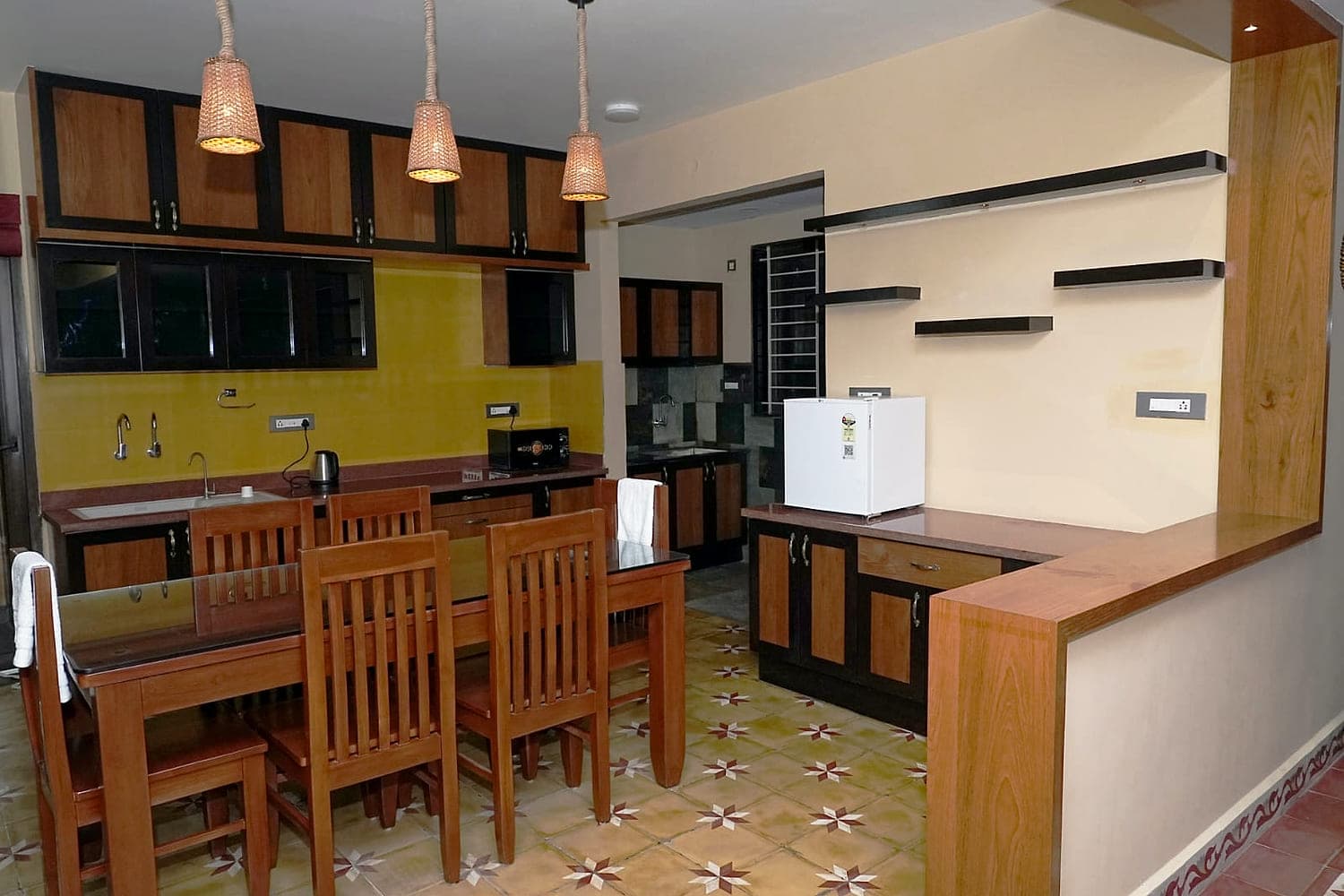 A dining table and kitchen in wood and yellow, on a patterned tiled floor.