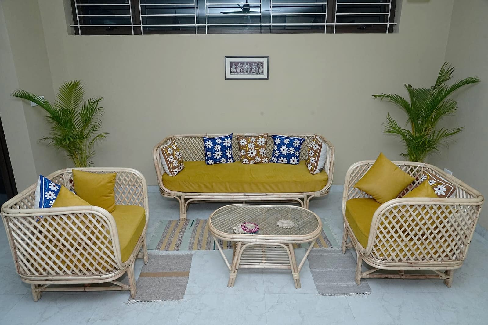 A living room furnished in cane, with two palms and a framed drawing on a plain wall.