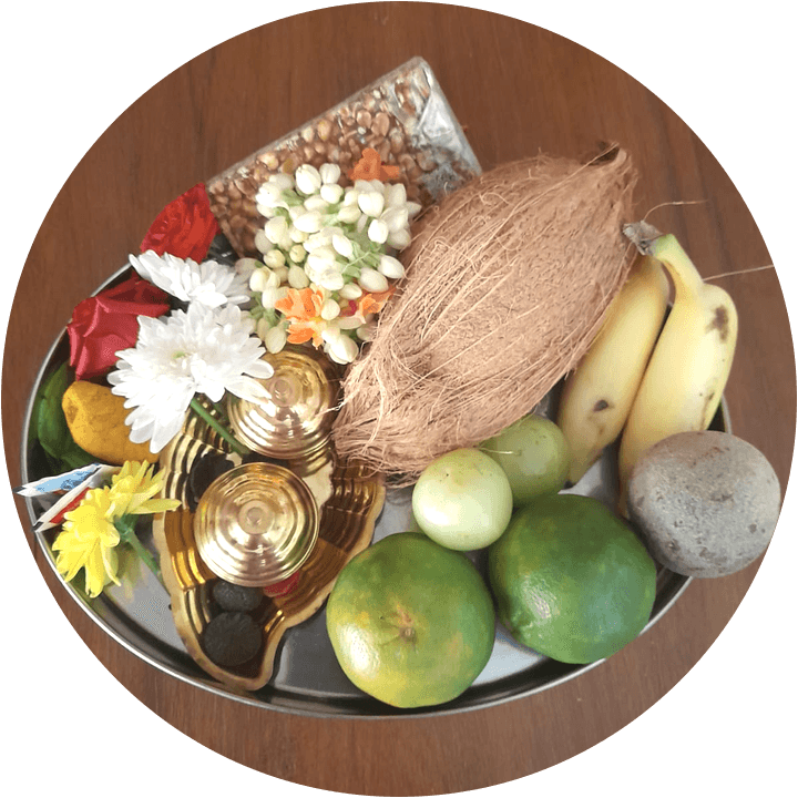 A taamboolam tray: jasmine and chrysanthemums, a coconut, bananas, limes, brass lamps and a grinding stone, arranged on a steel plate.